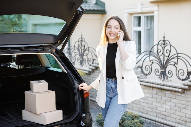 Joven mujer de negocios de pie en el auto con paquetes que llegan a casa en auto La chica está de pie y hablando por teléfono móvil cerca del auto Concepto de comprar productos en línea y entregarlos a casa