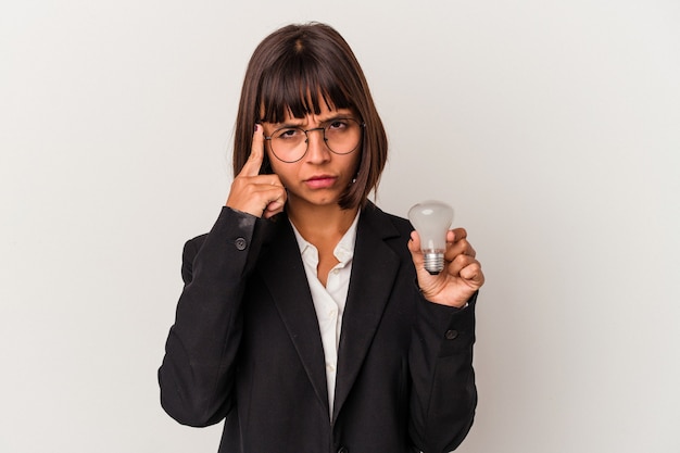 Joven mujer de negocios de raza mixta sosteniendo una bombilla aislada sobre fondo blanco señalando la sien con el dedo, pensando, centrado en una tarea.