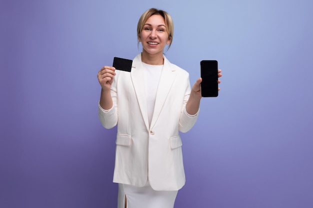 Joven mujer de negocios rubia europea con una chaqueta blanca y un vestido con una maqueta de tarjeta y teléfono