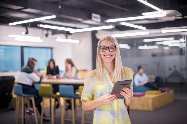 joven mujer de negocios rubia que trabaja en línea usando una tableta digital mientras está de pie en una oficina de inicio moderna con un equipo de negocios multiétnico en segundo plano