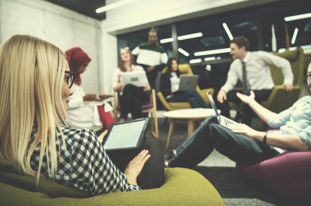 joven mujer de negocios rubia que trabaja en línea usando una tableta digital mientras está sentada en una oficina de inicio moderna con un equipo de negocios multiétnico en segundo plano