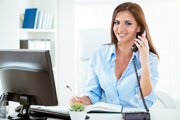 Foto joven mujer de negocios sexy sonriente sentada en la oficina frente al monitor y usando el teléfono. mirando a la cámara.