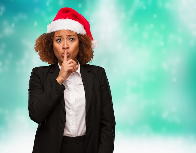 Joven mujer de negocios con un sombrero de santa chirstmas guardando un secreto o pidiendo silencio