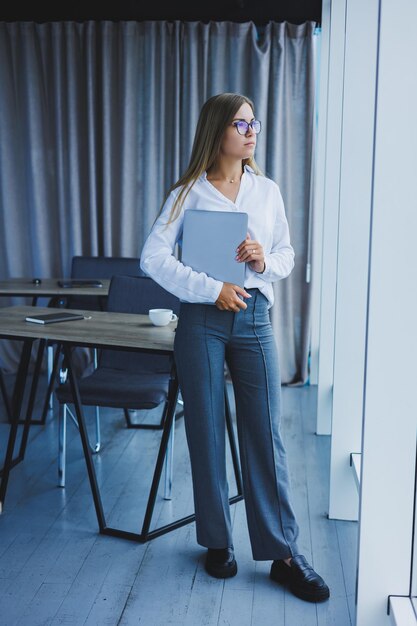 Una joven mujer de negocios sonriente está parada en la oficina junto a la ventana con una computadora portátil en las manos Gerente de mujer con gafas y camisa blanca