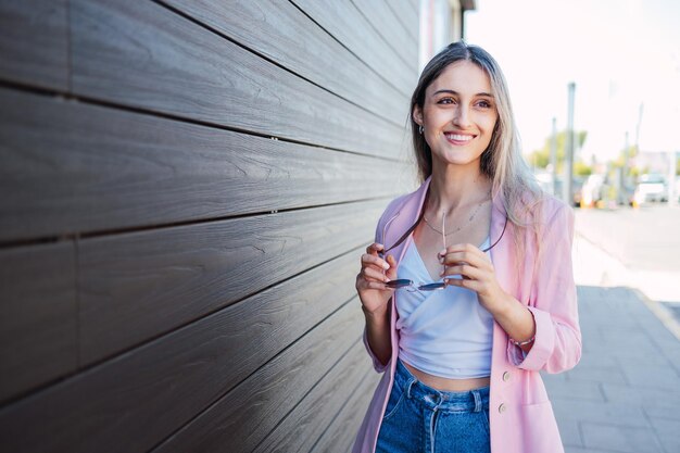 joven, mujer de negocios, sonriente, y, posición, al lado de, un, pared de madera, tenencia, gafas de sol