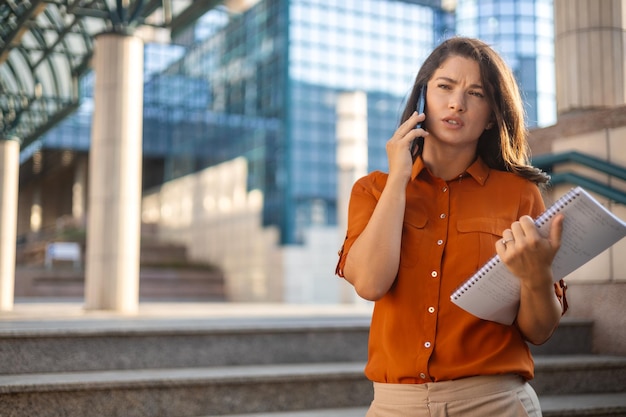 Una joven mujer de negocios sonriente con traje de pie en una escalera mecánica urbana usando aplicaciones en el teléfono celular leyendo noticias en la conexión rápida del teléfono inteligente revisando aplicaciones móviles al aire libre