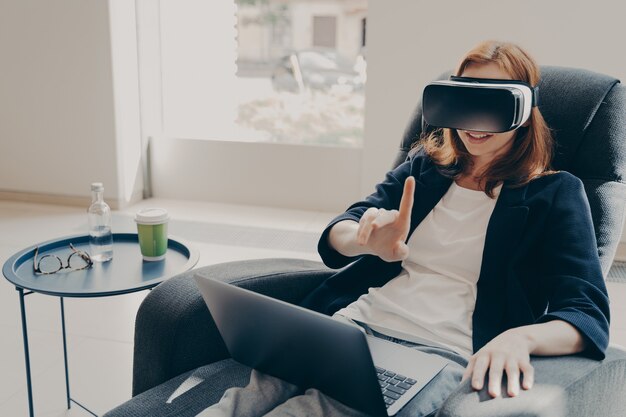 Joven mujer de negocios sorprendida feliz en auriculares VR tratando de tocar algo, usando la realidad virtual para los negocios, sentada en su acogedora oficina en casa, autónoma probando gafas mientras trabaja de forma remota