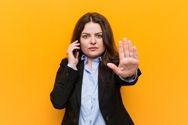 Joven mujer de negocios de talla grande con curvas que sostiene un teléfono de pie con la mano extendida que muestra la señal de stop, impidiéndole.