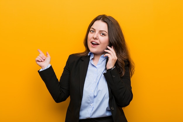 Joven mujer de negocios de talla grande con curvas sosteniendo un teléfono sonriendo alegremente señalando con el dedo lejos.