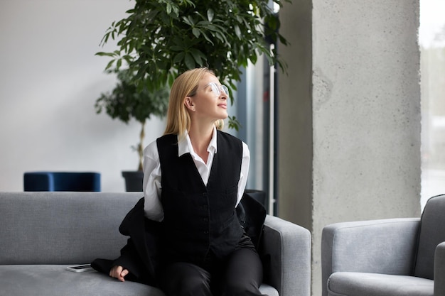 Joven mujer de negocios con traje sentada en el elegante sofá gris en un salón de negocios con plantas de interior