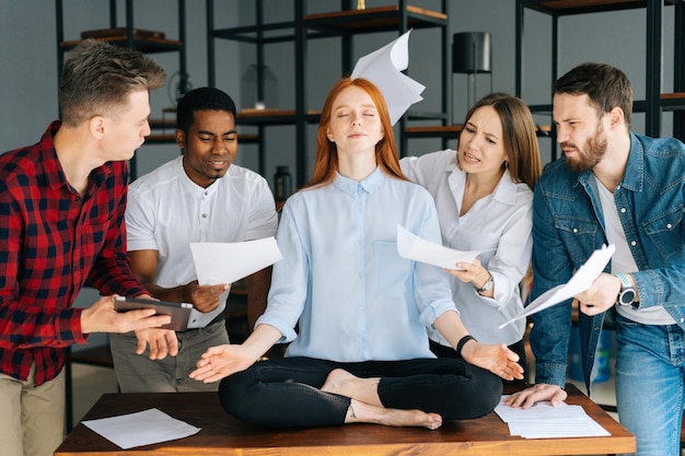 Una joven mujer de negocios tranquila medita en una reunión de negocios evitando la presión molesta a colegas enojados. Empresaria meditativa haciendo ejercicios de yoga para aliviar el estrés. Compañeros de trabajo sacudiendo documentos en papel.