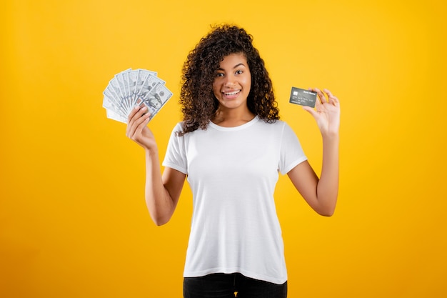 Joven mujer negra africana con dinero en dólares y tarjeta de crédito en mano aislado sobre amarillo