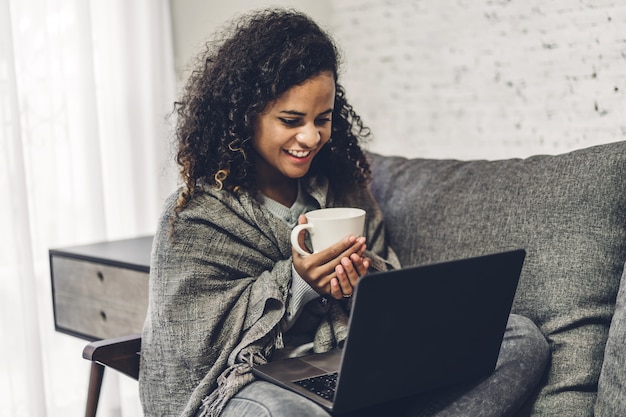 Joven mujer negra afroamericana relajarse y usar la computadora portátil. Mujer comprobando aplicaciones sociales y trabajando. Concepto de comunicación y tecnología