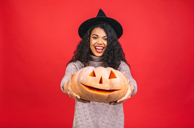 Foto joven mujer negra afroamericana sosteniendo una calabaza tallada aislado sobre fondo rojo.