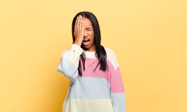 Joven mujer negra con aspecto somnoliento, aburrido y bostezando, con dolor de cabeza y una mano cubriendo la mitad de la cara