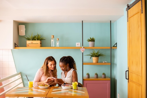 Joven mujer negra y caucásica mirando el teléfono móvil mientras bebe jugos frescos y desayuna saludablemente en la cafetería