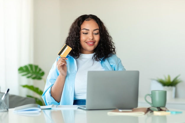Joven mujer negra de gran tamaño con tarjeta de crédito usando una computadora portátil en casa pidiendo comida o comprando espacio de copia en línea