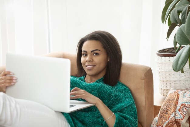 Foto joven mujer negra con ordenador portátil, sentado en la luminosa sala de estar.