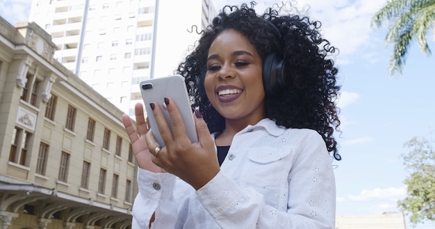 Joven mujer negra de pelo rizado caminando con teléfono celular. Enviar mensajes de texto en la calle. Gran ciudad.