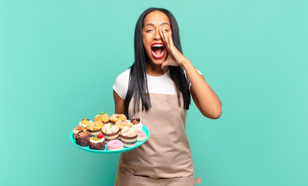 Joven mujer negra que se siente feliz, emocionada y positiva, dando un gran grito con las manos al lado de la boca, gritando. concepto de chef de panadería