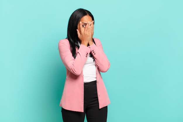 Joven mujer negra sintiéndose asustada o avergonzada, mirando o espiando con los ojos medio cubiertos con las manos. concepto de negocio