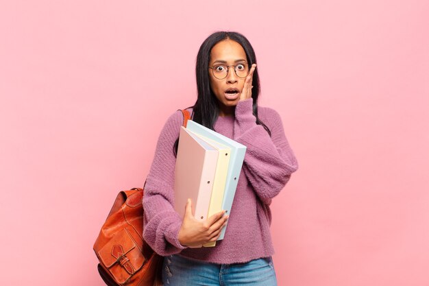 Joven mujer negra sintiéndose conmocionada y asustada, mirando aterrorizada con la boca abierta y las manos en las mejillas. concepto de estudiante