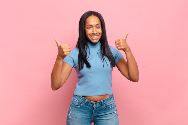 Joven mujer negra sonriendo con alegría y luciendo feliz, sintiéndose despreocupada y positiva con ambos pulgares hacia arriba