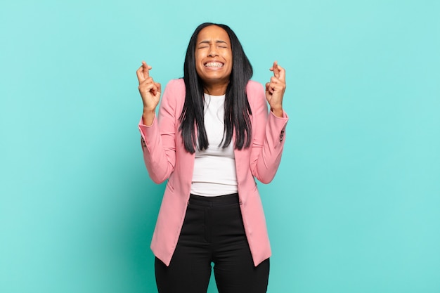 Joven mujer negra sonriendo y cruzando ansiosamente ambos dedos, sintiéndose preocupada y deseando o esperando buena suerte. concepto de negocio