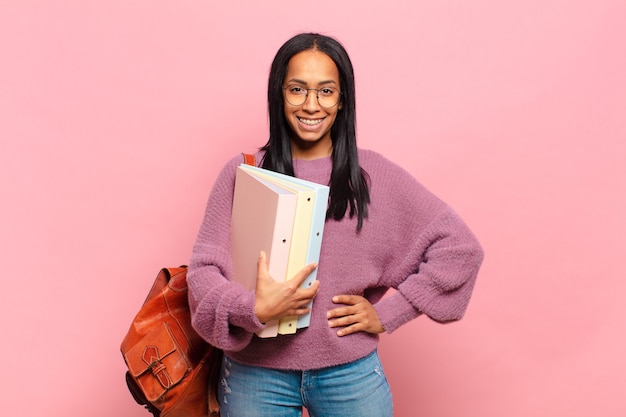 Joven mujer negra sonriendo felizmente con una mano en la cadera y actitud confiada, positiva, orgullosa y amistosa. concepto de estudiante