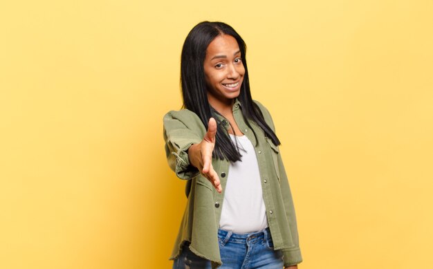 Joven mujer negra sonriendo, luciendo feliz, confiada y amigable, ofreciendo un apretón de manos para cerrar un trato, cooperando