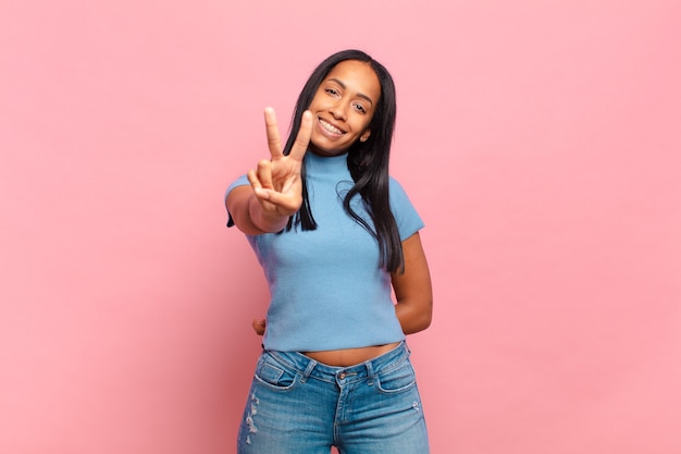 Joven mujer negra sonriendo y luciendo feliz, despreocupada y positiva, gesticulando victoria o paz con una mano