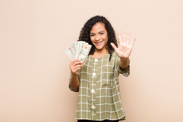 Joven mujer negra sonriendo y mirando amigable, mostrando el número cinco o quinto con la mano hacia adelante, contando con billetes de dólar