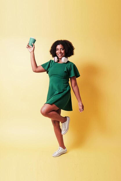 joven mujer negra sonriente con vestido verde con auriculares sosteniendo una taza de café