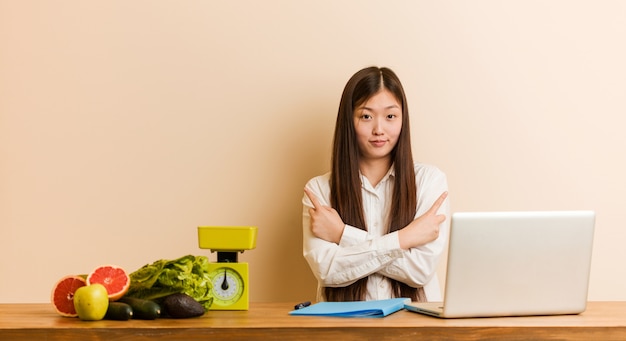 La joven mujer nutricionista china que trabaja con su portátil apunta hacia un lado, está tratando de elegir entre dos opciones.