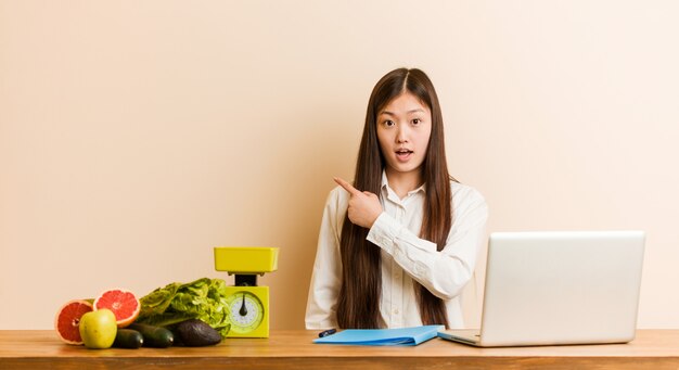 Joven mujer nutricionista china trabajando con su portátil apuntando hacia el lado