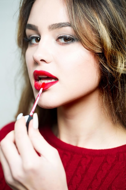 Joven mujer o niña muy sexy con cara linda y cabello largo y rubio poniendo lápiz labial rojo en los labios con pincel de maquillaje sobre fondo blanco