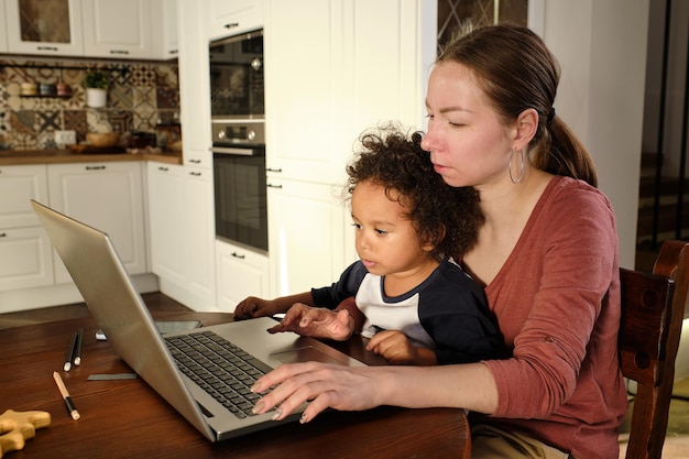 Joven mujer ocupada sosteniendo a su pequeño y lindo hijo mientras está sentado frente a la computadora portátil