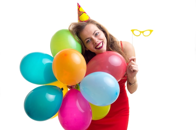 Joven mujer optimista con sombrero de celebración sosteniendo muchos globos de colores y palo de gafas