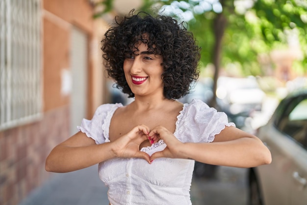 Joven mujer de oriente medio sonriendo confiada haciendo gesto de corazón con las manos en la calle