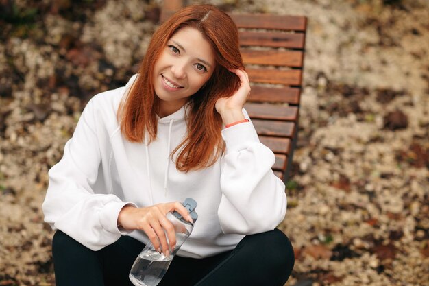 Joven mujer pelirroja deportiva sentada en un banco de madera en el parque con una botella de agua y mirando a la cámara mujeres sonrientes felices descansando después del entrenamiento