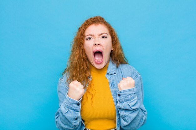 Joven mujer pelirroja gritando agresivamente con mirada molesta, frustrada, enojada y puños apretados, furiosa por la pared azul