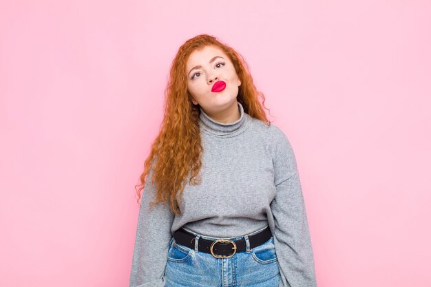 Joven mujer pelirroja que parece tonta y divertida con una expresión tonta, bromeando y bromeando contra la pared rosa