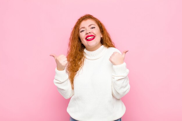 Joven mujer pelirroja sonriendo alegremente y luciendo feliz, sintiéndose despreocupada y positiva con ambos pulgares sobre la pared rosa