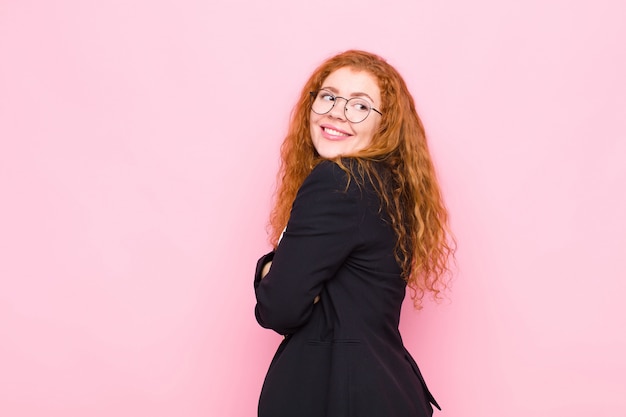 Joven mujer pelirroja sonriendo alegremente, sintiéndose feliz, satisfecha y relajada, con los brazos cruzados y mirando a un lado contra la pared rosa