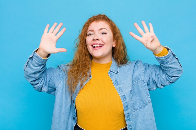 Joven mujer pelirroja sonriendo y mirando amigable, mostrando el número nueve o noveno con la mano hacia adelante, contando contra la pared azul