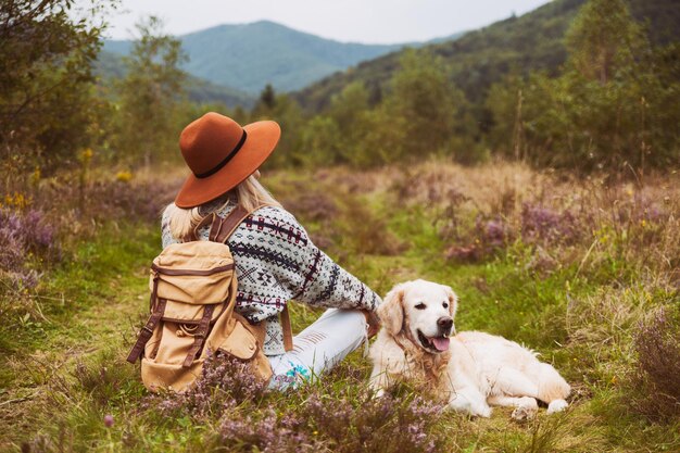 Joven mujer de pelo rubio hippie en suéter de lana con mochila textil y sonriente feliz perro golden retriever sentado en la hierba en el valle de las montañas de verano. Concepto de actividades de fin de semana y senderismo de mascotas.