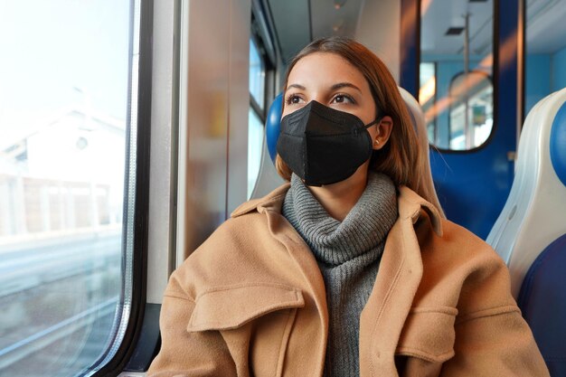 Joven mujer pensativa con mascarilla médica negra en el tren mirando por la ventana. Concepto de viajar y usar el transporte público durante la pandemia.