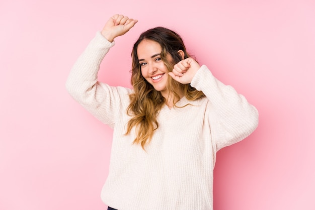 Joven mujer posando en una pared rosa celebrando un día especial, salta y levanta los brazos con energía.