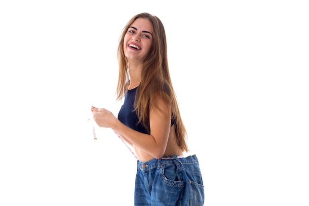 Joven mujer positiva con cabello castaño largo sosteniendo sus jeans de tamaño mucho más grande con centímetro sobre fondo blanco en el estudio