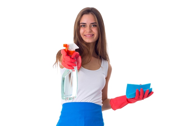 Joven mujer positiva con camisa blanca y delantal azul con guantes rojos sosteniendo plumero y detergente sobre fondo blanco en el estudio